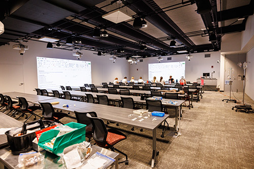 A classroom featuring tables and chairs arranged in front of a large projector screen, ready for a presentation.