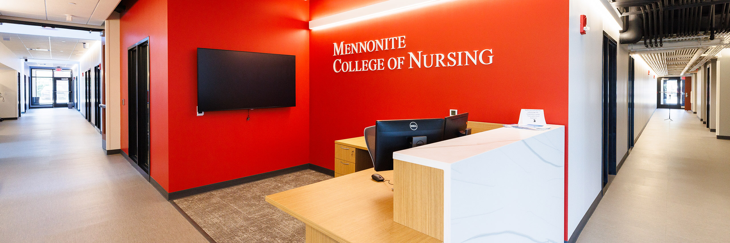 Reception desk at Mennonite College of Nursing, with a red wall featuring the college name. A hallway is visible to the left.